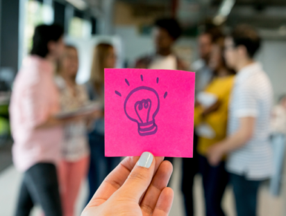 A post-it with a light bulb as a symbol for innovation. In the background you can see a group of employees working on these innovations. Illustration of the article "Let's Rock and Roll: How to Innovate Sustainably in Bureaucratic Organisations?" by Fernanda Arreola (ESSCA) for European Business Review.