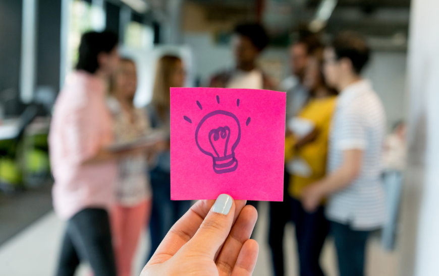 A post-it with a light bulb as a symbol for innovation. In the background you can see a group of employees working on these innovations. Illustration of the article "Let's Rock and Roll: How to Innovate Sustainably in Bureaucratic Organisations?" by Fernanda Arreola (ESSCA) for European Business Review.